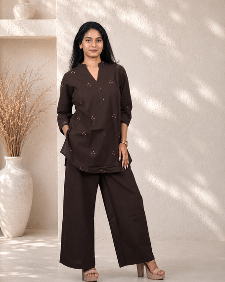 Woman wearing a brown outfit standing in a room with a white wall and decorative vase.